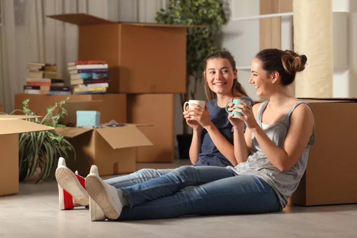 Two girls sitting on floor in housing talking, there are moving boxes around them