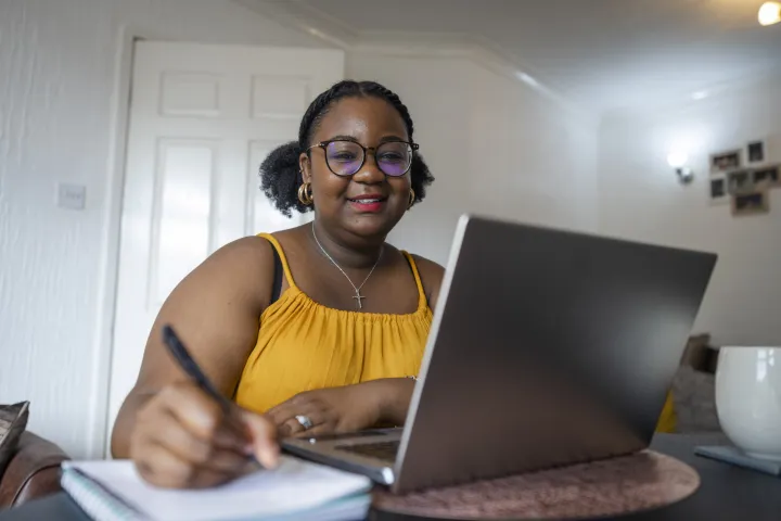 Woman in a yellow top taking notes by a laptop